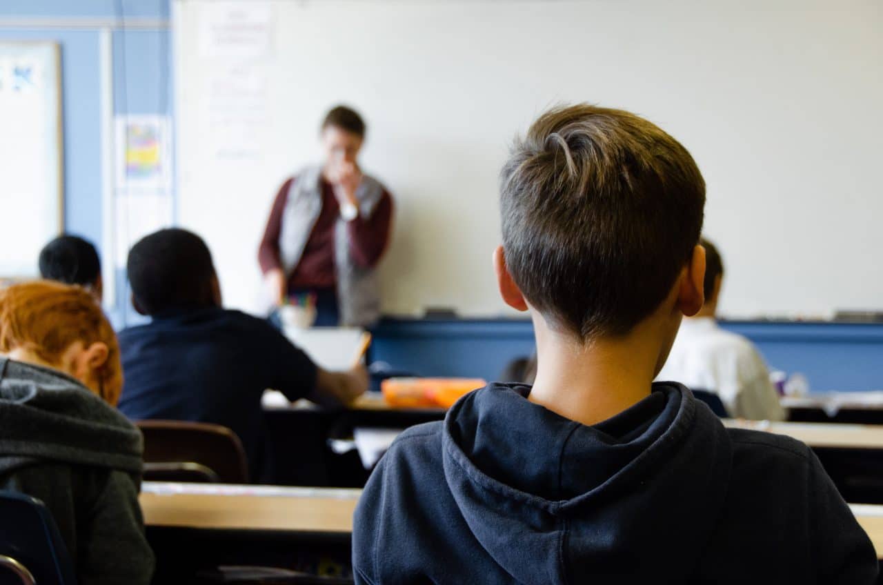 A teacher at the front of a classroom with students in the background.