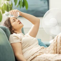 Young woman sitting on the couch at home and keeping a fresh water bottle on her forehead, she is cooling down from the heat.