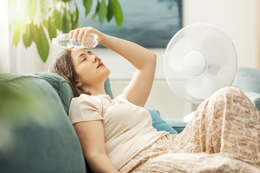 Young woman sitting on the couch at home and keeping a fresh water bottle on her forehead, she is cooling down from the heat.